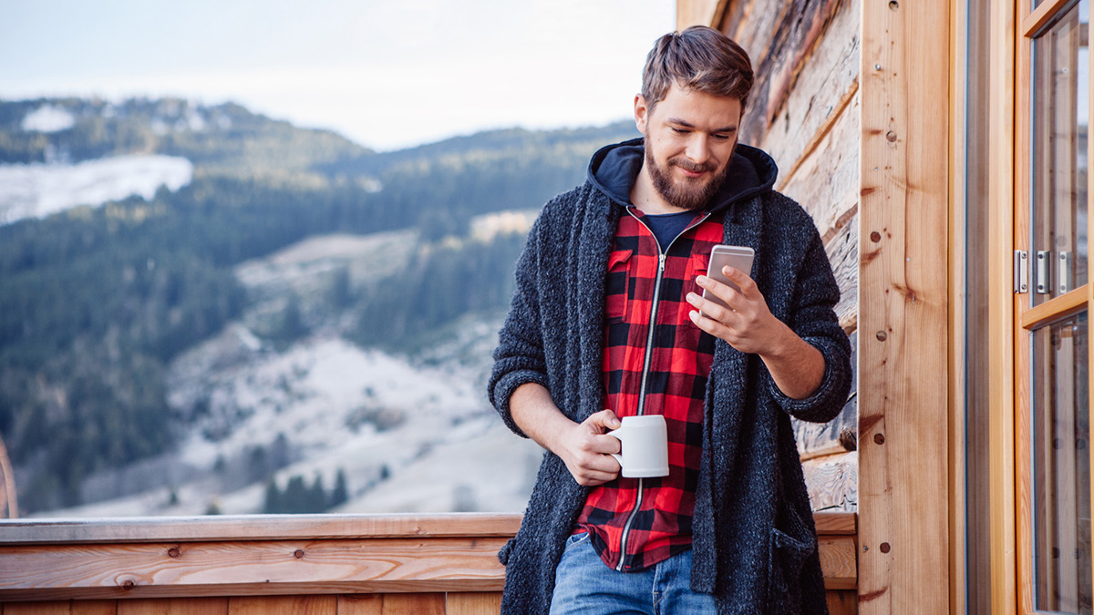 Mann på hytteterrassen med kaffe og mobiltelefon. Foto.